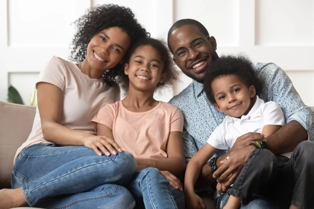 Portrait of happy young african American family with little kids sit relax on couch cuddling, smiling black parents rest on sofa hug preschooler children posing for picture at home together