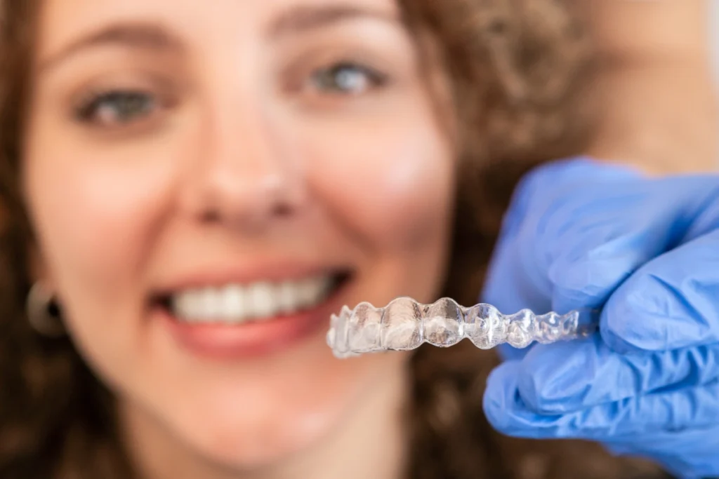 Woman smiling while holding clear Invisalign aligner tray in gloved hands.